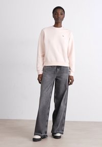 Világos rózsaszín pulóver, kerek nyakkivágással, bordázott mandzsettával és szegéllyel. Széles lábú szürke farmerral és tömör fekete-fehér cipővel párosítva.