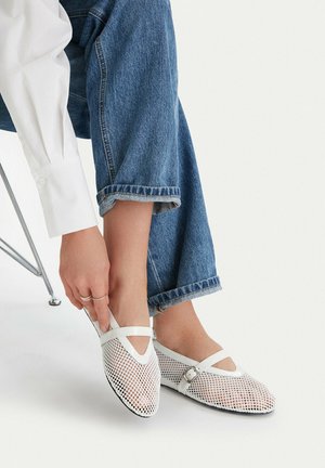 Osoba oblečená v modrých džínách s ohrnutými nohavicemi a bílém tričku, zapínající nastavitelné bílé síťované tenisky se zapínáním na přezky.