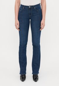 Tamno plave flared traperice s srednjim strukom, prednjim džepovima i detaljima šivanja, u kombinaciji s crnim cipelama s oštrim nosom.