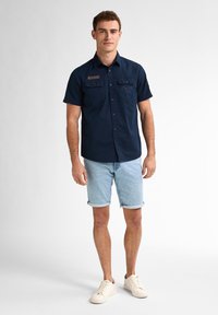 Camicia blu navy a maniche corte con due tasche sul petto, abbinata a pantaloni corti in denim azzurro arrotolati e sneakers beige.