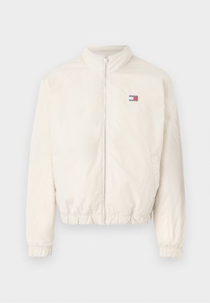 Veste bomber légère en crème avec fermeture éclair à l'avant, poignets élastiques et un petit logo en rouge et bleu. Texture lisse, design décontracté.