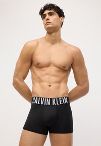 Crne bokserice s bijelim logotipom "CALVIN KLEIN" na pojasu. Glatka tekstura, pripijeni dizajn koji ističe tijelo.