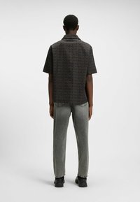 Camicia a maniche corte con colletto in tessuto scuro, decorata da un motivo con logo "HUGO" su tutta la superficie; indossata con jeans grigi e scarpe nere.