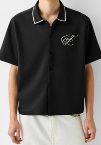 Czarna koszulka polo z krótkim rękawem, z zapięciem na guziki, z białym obszyciem na kołnierzu i haftowanym logo na lewym przedzie.