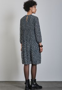 El vestido gris con estampado presenta diseños en espiral, mangas tres cuartos y una abertura en la parte posterior. Combinado con botas negras hasta el tobillo y medias.
