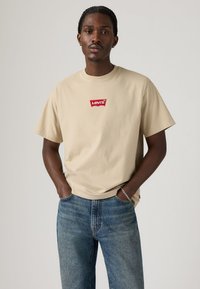 Beige bomull T-skjorte med en rød Levi's-logo-lapp. Har korte ermer og en avslappet passform, kombinert med blå denimbukser.