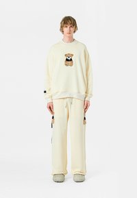 Conjunto de sudadera y pantalones crema con bordado de un oso en el pecho y las piernas, con detalles de cordón y tejido texturizado.