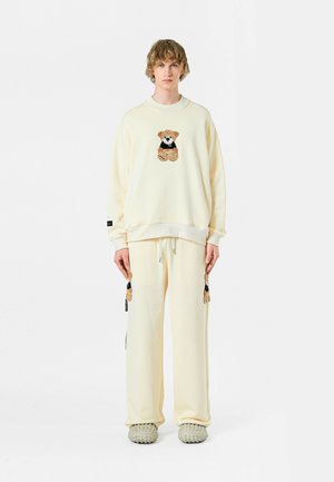 Conjunto de sudadera y pantalones crema con bordado de un oso en el pecho y las piernas, con detalles de cordón y tejido texturizado.