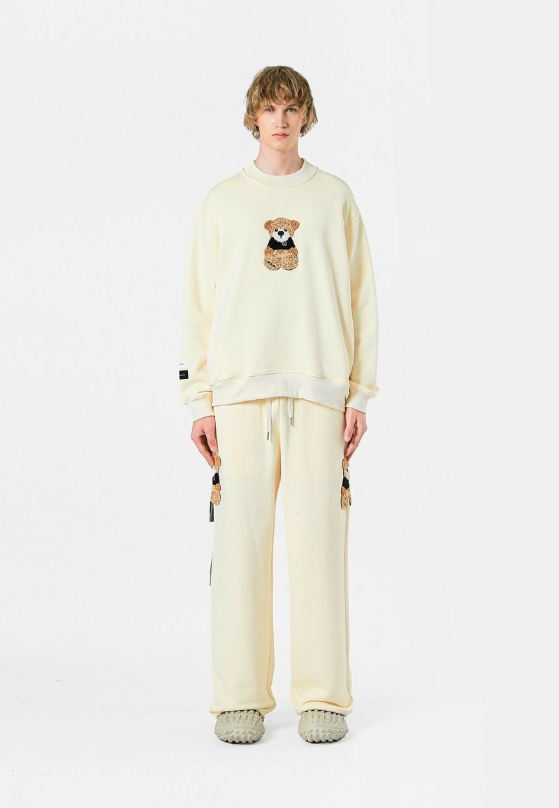 Conjunto de sudadera y pantalones crema con bordado de un oso en el pecho y las piernas, con detalles de cordón y tejido texturizado.