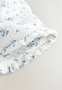 Bílá látka s modrým květinovým vzorem, s volánkovými okraji. Textura se jeví jako měkká a mírně zmačkaná, což naznačuje lehký a vzdušný design.