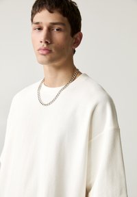 Pull en maille blanche à col rond et manches courtes, à la coupe décontractée, agrémenté d'un collier en chaîne argentée.