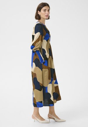 Žena stojící bokem, oblečená v dlouhých šatech s abstraktním vzorem v modré, hnědé, béžové a černé barvě, s béžovými nízkými pantoflemi na podpatku s otevřenou patou.