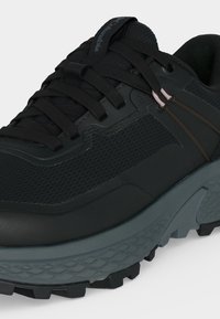 Chaussure de trail running noire et grise robuste avec empeigne en mesh, semelle épaisse texturée et lacets noirs, avec un logo de marque discret sur la languette.