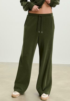 Personne portant un pantalon ample vert olive avec cordon et des baskets beige, levant un haut vert foncé pour dévoiler le ventre, debout sur un sol uni.
