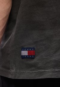 Κοντινό πλάνο σκούρου γκρι υφάσματος με ένα μικρό ορθογώνιο λογότυπο Tommy Jeans σε κόκκινο, άσπρο και μπλε κοντά στο τελείωμα.