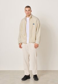 Mikina z flísu v krémovo béžovej farbe, so zipsom vpredu a bočnými vreckami. Dizajn obsahuje malý viacfarebný logo na hrudi.