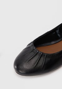Black leather ballet flat shoe with gathered detailing on the rounded toe and brown cushioned insole on a white background.