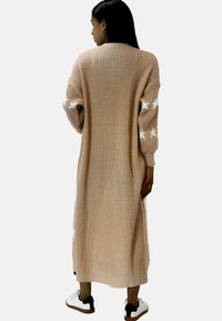 Larga cardigan de punto beige con patrones de estrellas blancas en las mangas, con un ajuste holgado y puños acanalados. Combinada con zapatillas blancas.