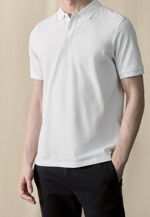 Homme portant un polo blanc uni à manches courtes avec col et un pantalon noir, debout avec une main dans la poche sur un fond neutre.
