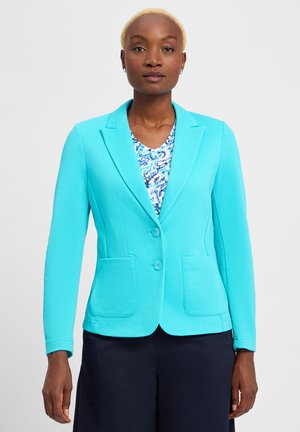 Frau mit kurzen blonden Haaren, die ein tailliertes türkisfarbenes Sakko über einem blau gemusterten Oberteil und dunklen Hosen trägt, steht vor einem schlichten Hintergrund.