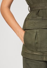 Gilet utilitaire en coton vert olive avec une finition texturée, présentant une taille cintrée, de grandes poches avant et des détails cousus.
