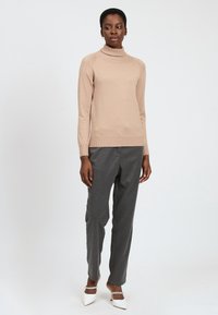 Maglione beige a collo alto con orlo a coste, abbinato a pantaloni grigi su misura e mule bianche con tacco. Il maglione ha una leggera texture luccicante.