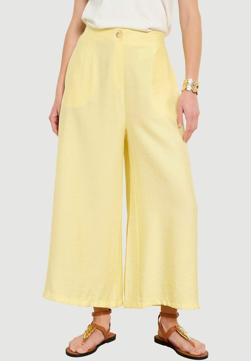 Mujer con pantalones amarillos de talle alto y pierna ancha, top blanco sin mangas, pulsera dorada y sandalias planas marrones con adornos dorados.