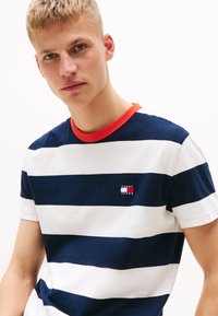 Navy og hvit stripete bomull T-skjorte med korte ermer og rød krage. Har en liten "Tommy Jeans"-logo på brystet.