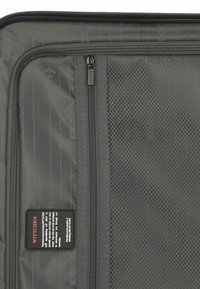 Gray travel bag interior featuring textured mesh pocket, zippers, and subtle airplane graphic pattern on smooth surface. Brand label visible.