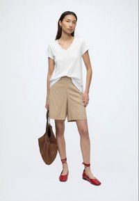 Femme portant un t-shirt blanc à col en V, un short beige longueur genou, des ballerines rouges à nouer à la cheville, et tenant un grand sac à main marron.