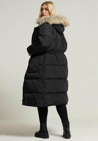 Czarny, długi płaszcz puchowy z kapturem z sztucznego futra, o pikowanej strukturze i kieszeniach bocznych. Noszony z czarnymi legginsami i botkami za kostkę.