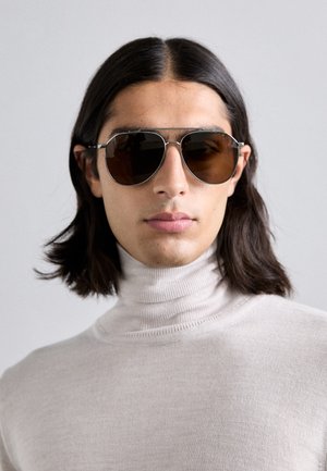 Gafas de sol con lentes de color marrón, montura de metal plateado y forma clásica de aviador, llevadas por una persona con un suéter de cuello alto beige claro.