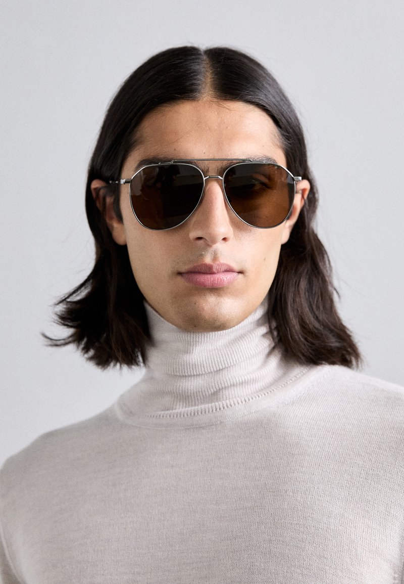 Saulesbrilles ar brūnām tonētām brillēm, sudraba metāla rāmjiem un klasisku aviatoru formu, kuras valkā persona gaiši bežā turtleneck džemperī.