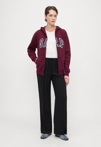 Sudadera burdeos con logo "GAP", camiseta blanca debajo, pantalones amplios de pierna ancha plisados en negro y zapatillas deportivas grises. Tejido suave, corte relajado.
