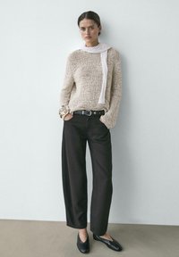 Kvinde stående med hænderne i lommerne, iført beige striktrøje, hvidt tørklæde, sorte bukser med vide ben, sorte sko og guldbøjler.