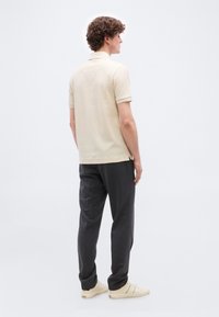 Άνδρας με σγουρά μαλλιά που φοράει κρεμ πόλο με υφή, σκούρο γκρι παντελόνι και κρεμ αθλητικά παπούτσια, στέκεται στραμμένος ελαφρώς δεξιά μπροστά σε απλό φόντο.