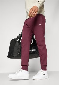 Jogging en maroon avec des poignets élastiques, écusson logo sur la jambe, associés à des baskets blanches. L'individu tient un sac de sport noir avec du texte blanc.