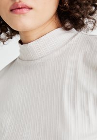 Hellbeige, gerippter Rollkragenpullover mit hohem Kragen. Der Stoff hat eine glatte Textur und dezente vertikale Linien für zusätzliche Details.