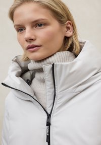 Chaqueta aislante de color gris claro con cuello alto, cremallera frontal y herrajes negros. Lleva puesta sobre un jersey de cuello alto beige de punto acanalado.