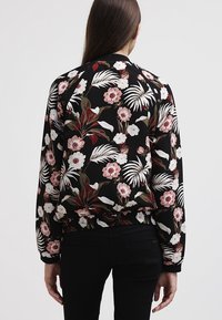 Kvinde vendt væk iført en sort bomberjakke med et pink og hvidt blomster- og bladmønster, parret med sorte bukser.