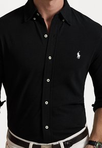 Černá košile s dlouhým rukávem na knoflíky s ostrým límečkem. Na texturované látce je malé bílé vyšívané logo a kontrastní bílé knoflíky.