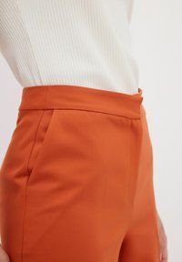 Oranje op maat gemaakte broek met een gladde textuur, hoge tailleband en zijzakken. Gecombineerd met een witte geribbelde top met lange mouwen.