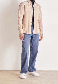 Beżowa koszula z długim rękawem zapinana na guziki, pod spodem czarny golf, do tego jasnoniebieskie dżinsy i białe trampki.