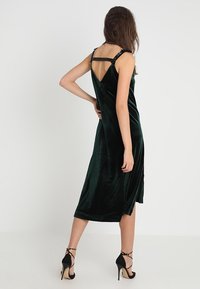 Samtgrünes Kleid mit V-Ausschnitt, midi-lange Schnittform und dünnen Trägern. Kombiniert mit schwarzen High-Heelsandalen mit Kettenakzenten.
