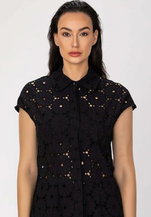 Femme aux longs cheveux foncés porte un chemisier noir en dentelle ajourée à motifs floraux, manches courtes, avec un col, sur un fond clair uni.