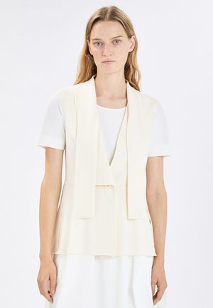 Donna con capelli biondi lisci che indossa un gilet crema senza maniche sopra una camicia bianca a maniche corte, in piedi davanti a uno sfondo chiaro e uniforme.