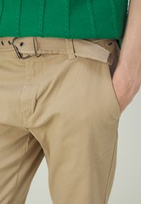 Kaki hlače s opuštenim krojem, koje imaju bež platneni pojas. Tkanina je glatka, s bočnim džepovima i suptilnim detaljima šivanja.