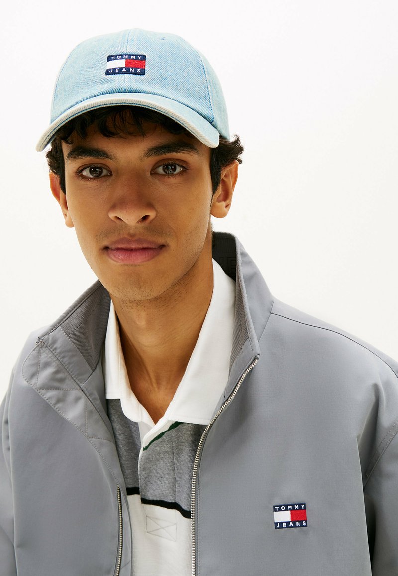 Jonge man met een lichtblauwe Tommy Jeans pet, een grijze jas met het Tommy Jeans-logo en een wit-grijs overhemd met kraag.