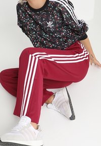 Černý top s květinovým vzorem od Adidas s dlouhými rukávy, červené teplákové kalhoty s bílými pruhy a bílé sportovní boty s texturovanou podrážkou.