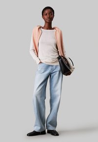 Top beige chiaro a maniche lunghe con sovrapposizione senza maniche rosa, abbinato a jeans larghi blu chiaro e scarpe nere, mentre tiene una borsa nera.